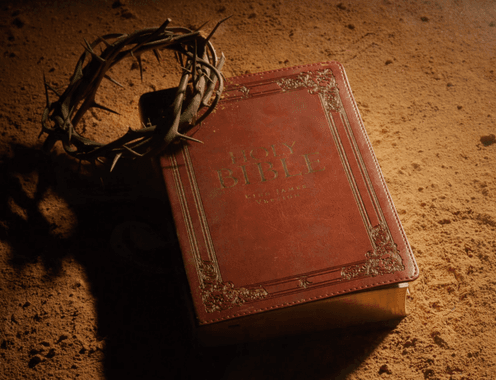 A leather-bound Holy Bible resting on sand beside a crown of thorns, lit dramatically