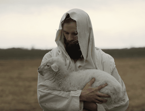 Jesus, portrayed as a shepherd in traditional robes, tenderly holding a lamb in an open field
